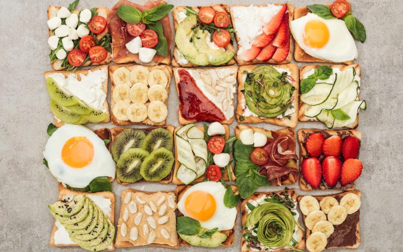 Top view of toasts with fried eggs, cut vegetables and fruits on textured surface